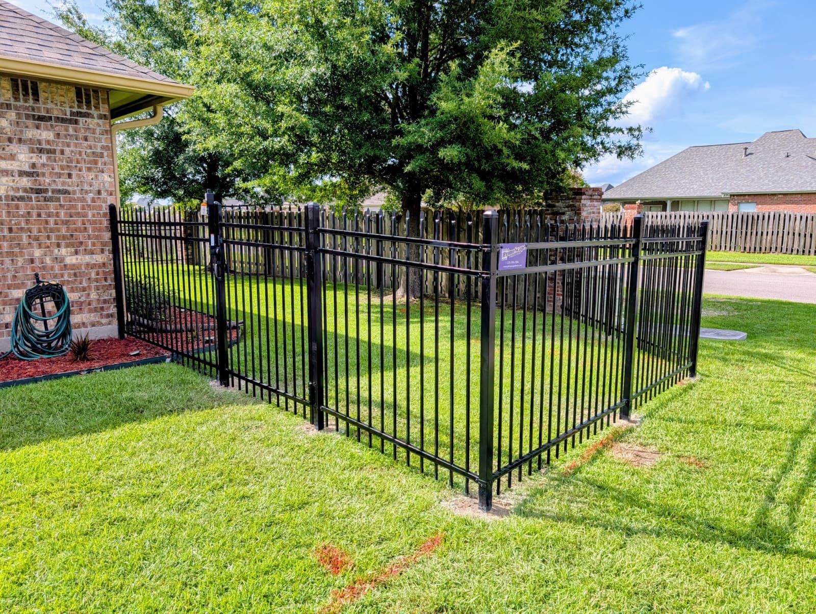 Black metal fence enclosing a grassy backyard beside a brick house, with a tree and neighbor homes beyond.