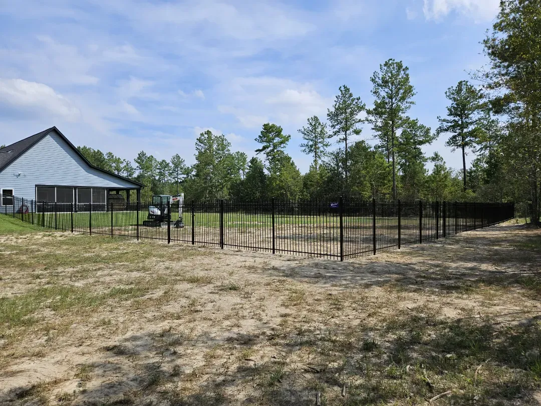 Fenced grassy lot beside a house under construction, with trees and a partly cloudy sky.