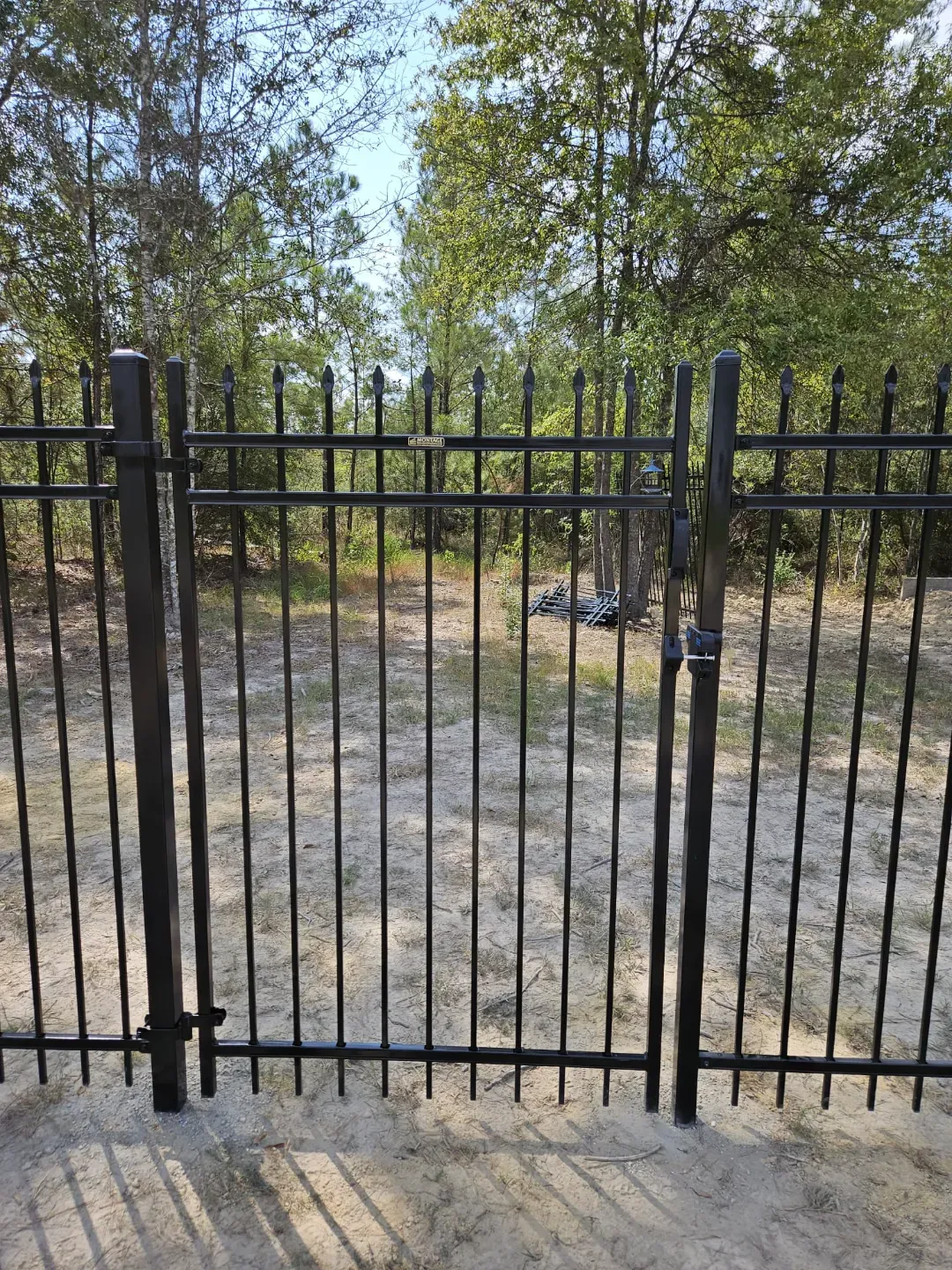Black metal gate opening to a wooded path with dry grass and trees beyond.