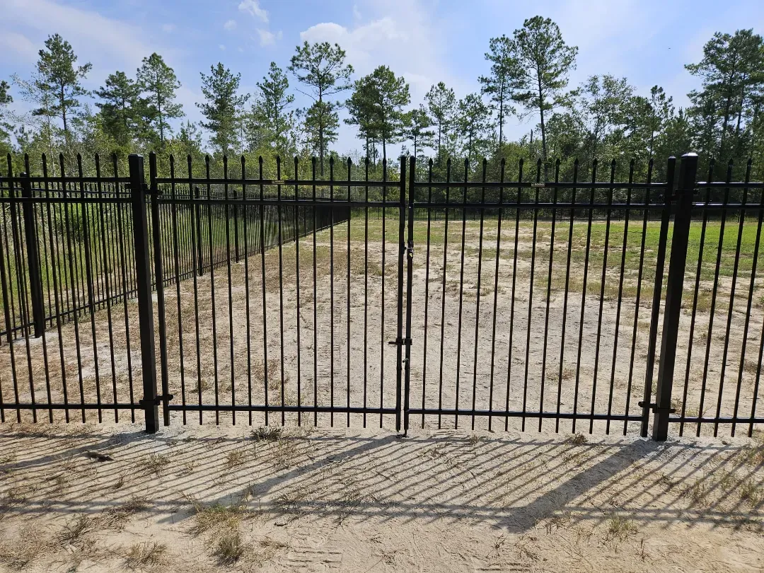 Black metal fence with a gated entrance along a dirt path in a wooded area