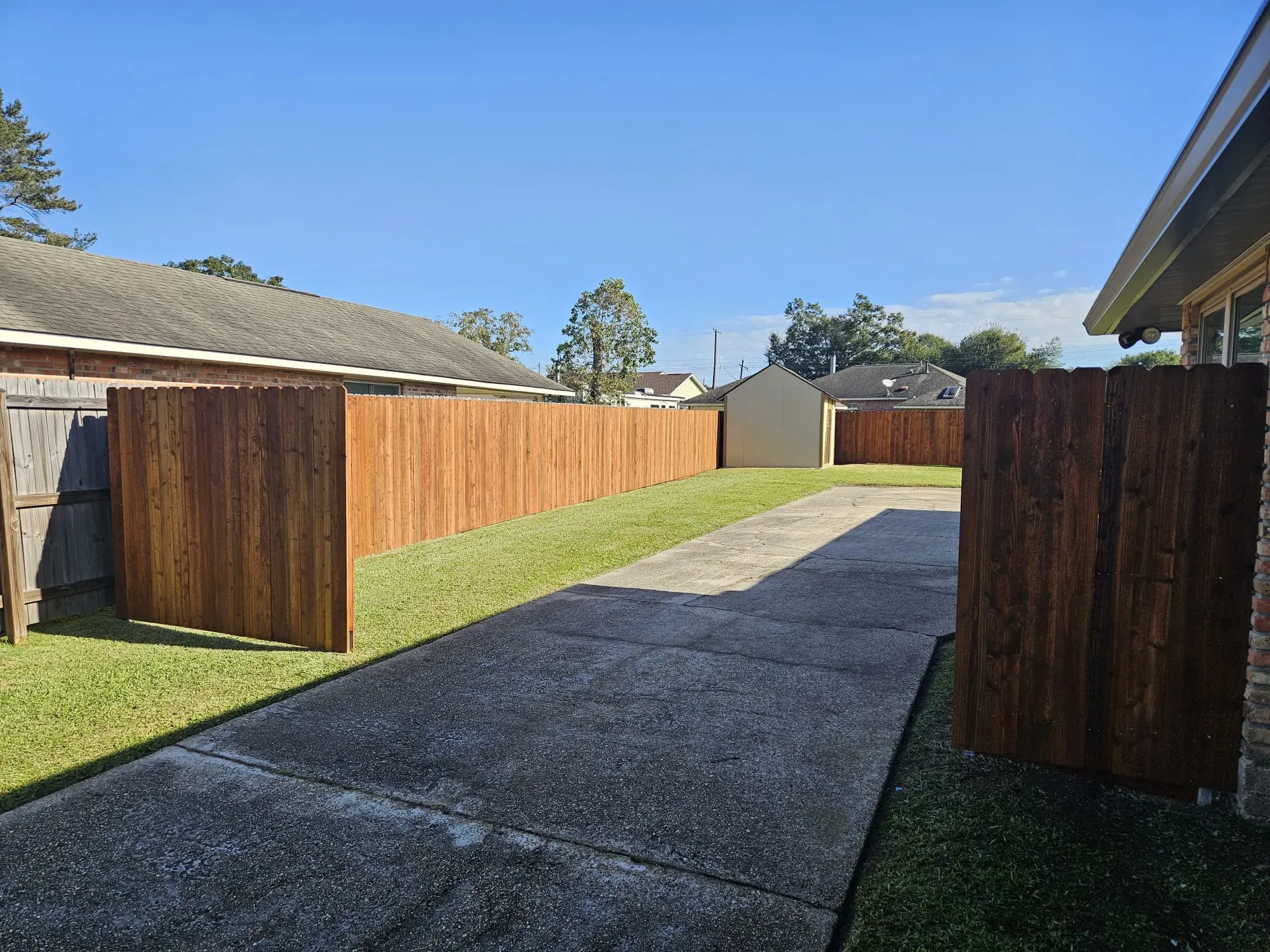 Narrow concrete driveway between wooden fences leading to a garage under a blue sky.