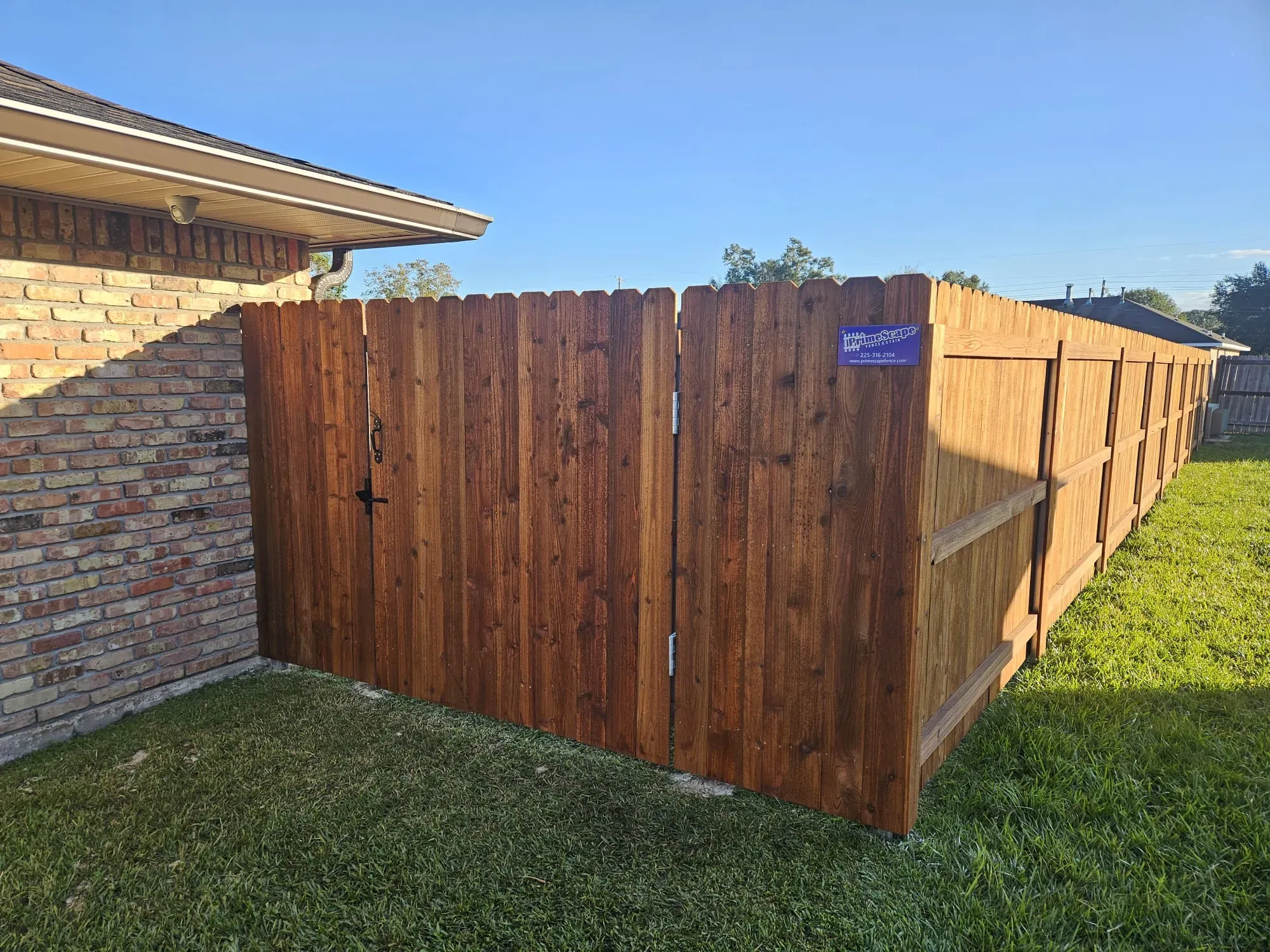 Wooden privacy fence beside a brick house on a grassy yard under a clear blue sky