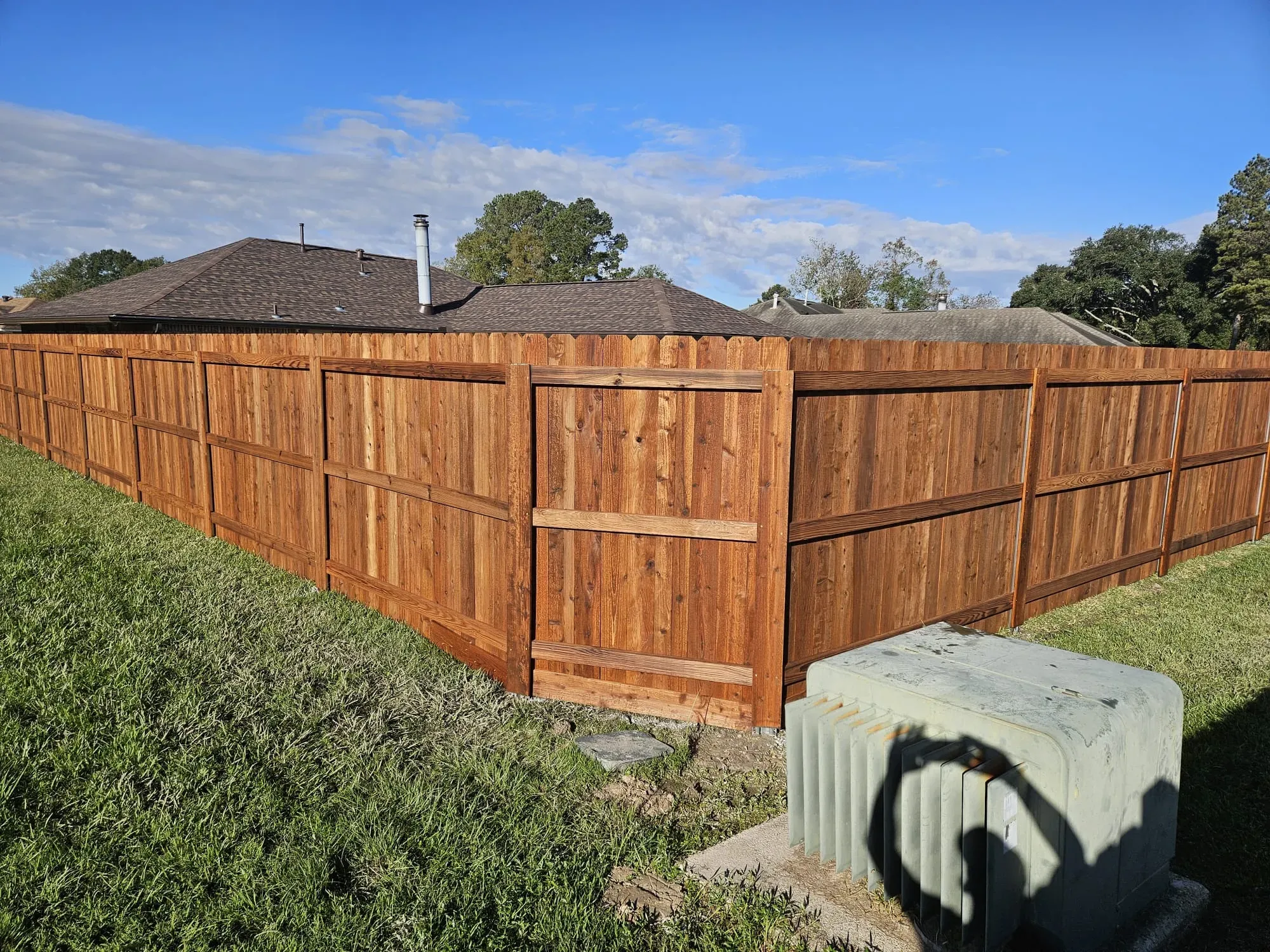 Wooden privacy fence around a yard on a sunny day, with green shrubs and a utility box in front.
