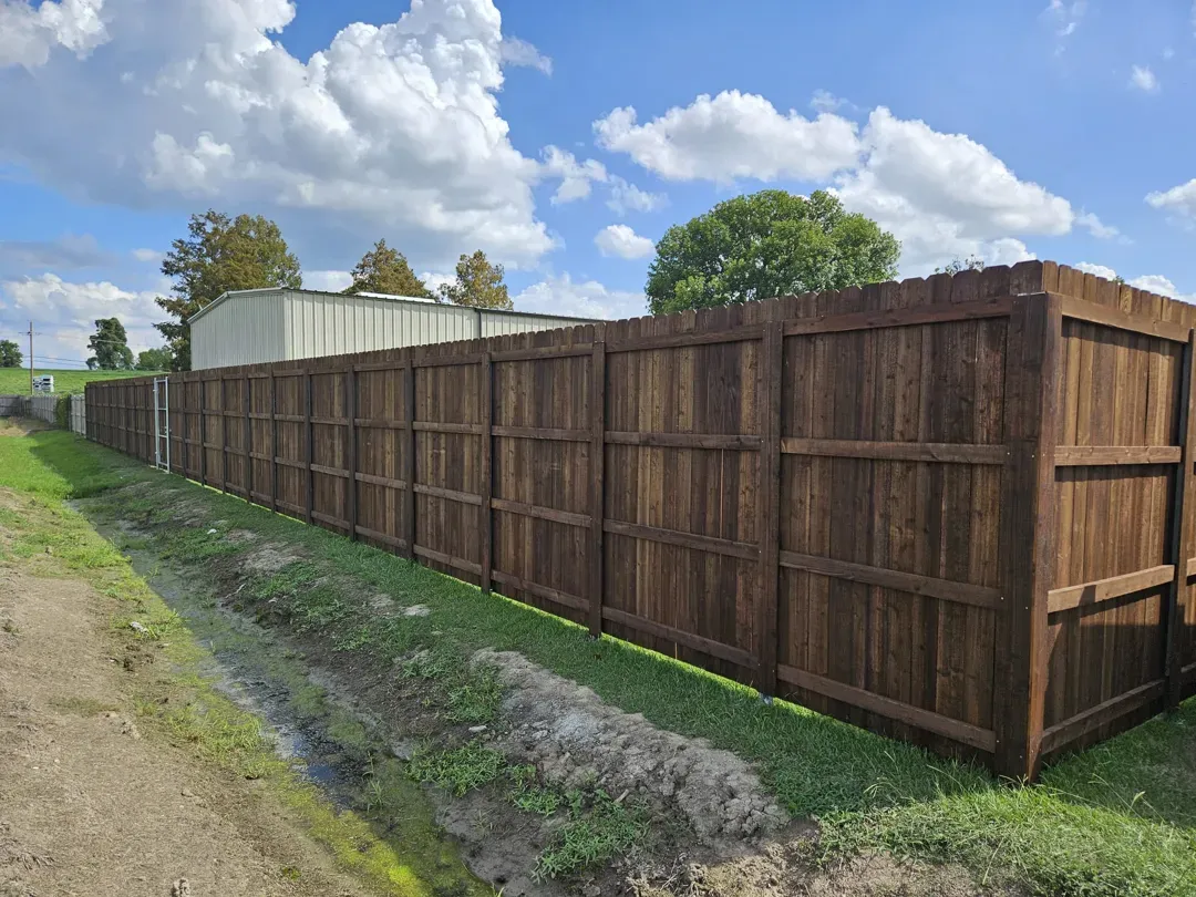 Long brown wooden privacy fence beside a muddy dirt path under a blue sky with clouds