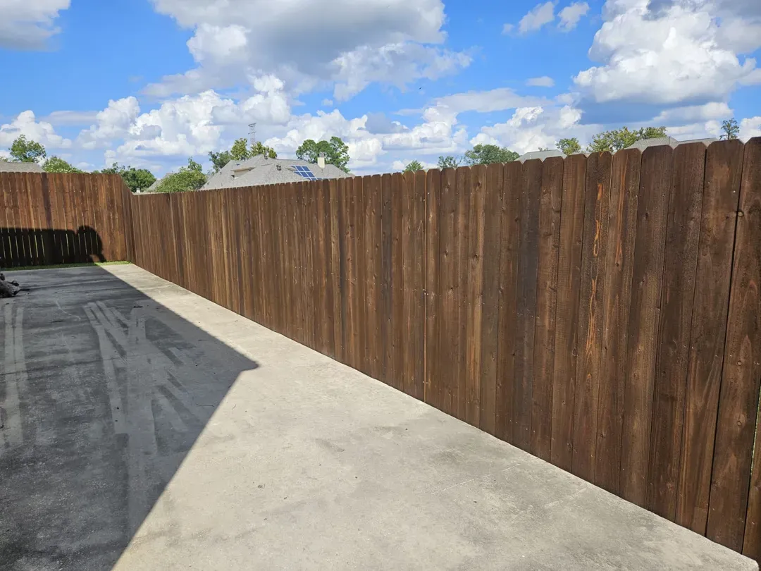 Concrete driveway beside a tall wooden fence under a blue sky with clouds