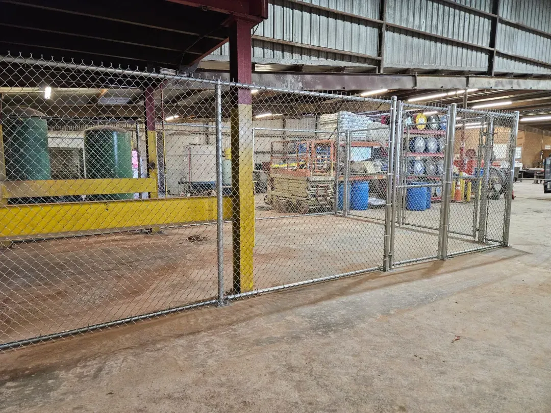 Chain-link gate at an industrial warehouse entrance, with blue barrels and stacked goods inside.
