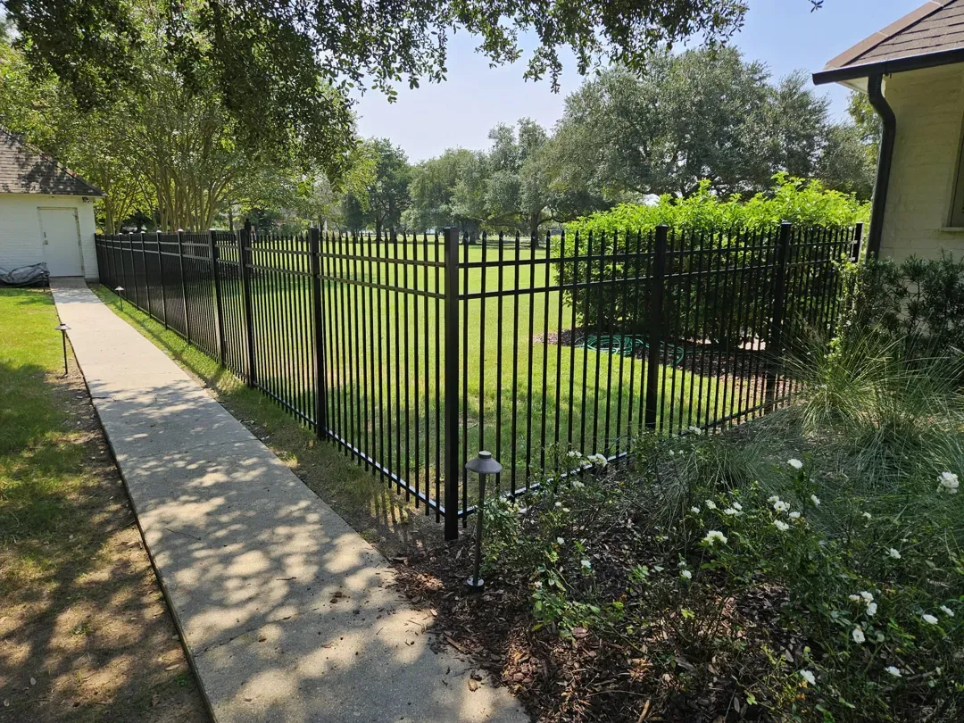 Black metal fence beside a concrete sidewalk, bordering a green lawn and trees on a sunny day