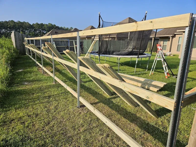 Wooden fence framework leaning along a grassy yard beside a house and ladder