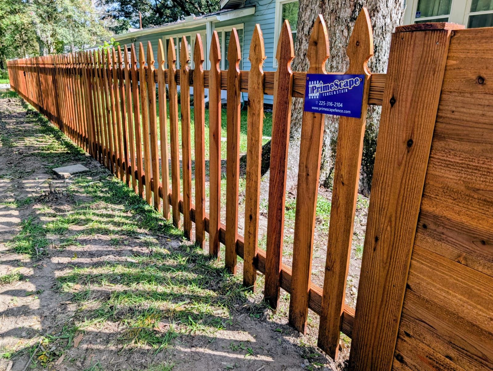 Wooden picket fence beside a gravel path, with trees and a blue sign in sunlight.