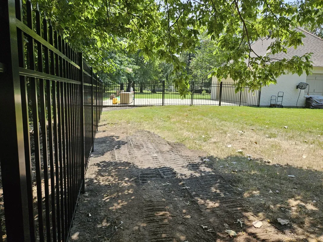 Shaded park path beside a black metal fence, leading to a sunlit grassy area and white building.