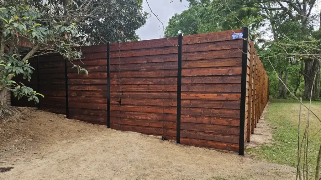 Wooden fence with black metal posts along a sandy path under trees in a rural area