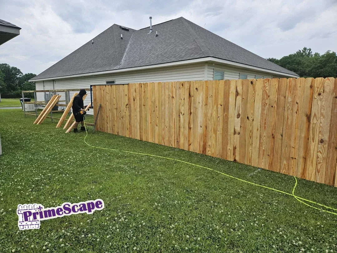 A worker installs a wooden privacy fence beside a gray-roofed house on a grassy lawn.