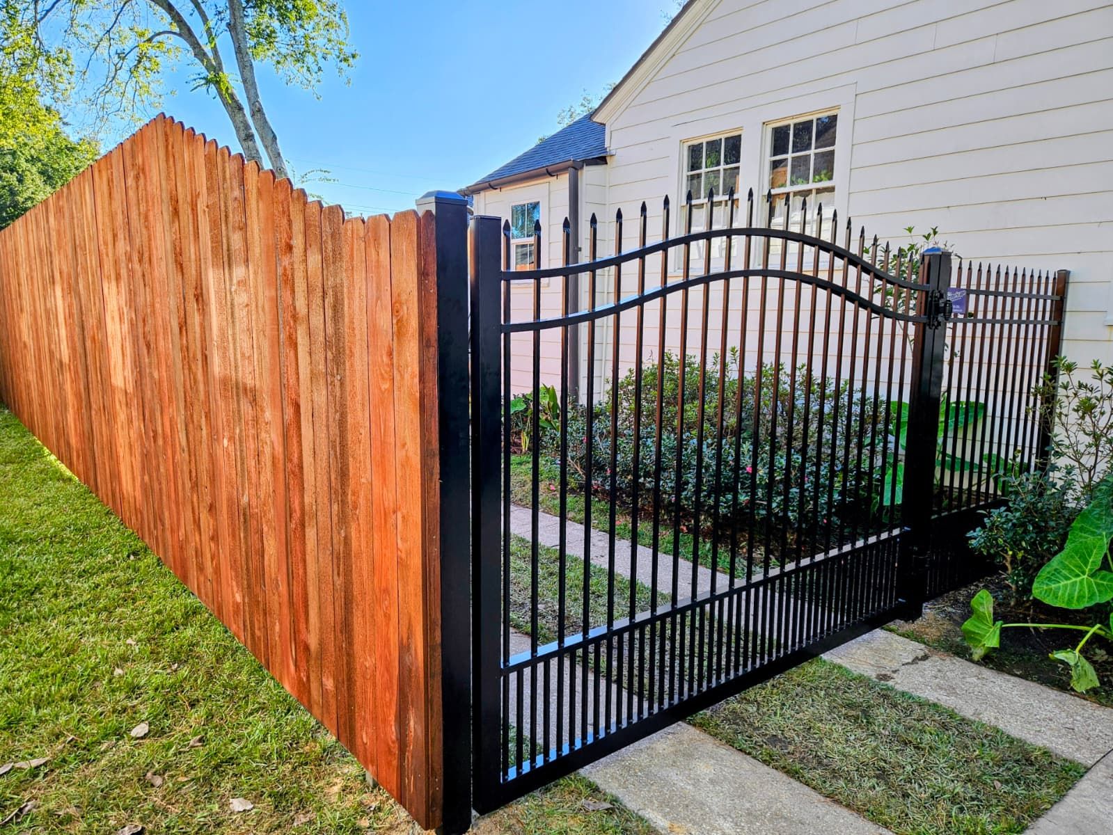 Black wrought-iron gate beside a wooden privacy fence in a front yard with a white house behind it