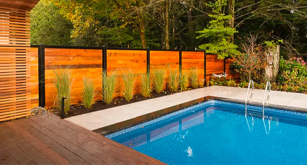Backyard swimming pool with blue water, wooden deck, orange privacy fence, and tall grasses