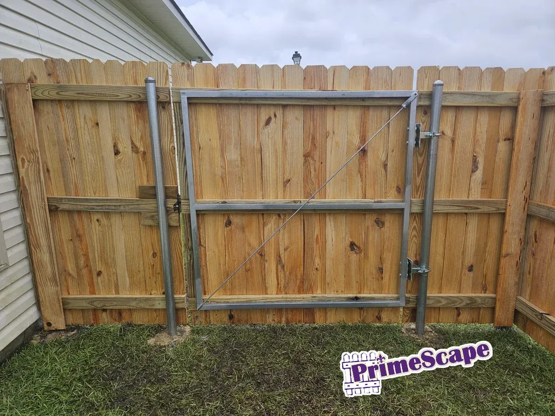 Wooden privacy fence with a metal-framed gate in a grassy backyard, PrimeScape sign at bottom right