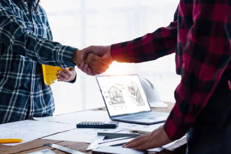 Two people shaking hands over a desk with a laptop and architectural sketchsheets