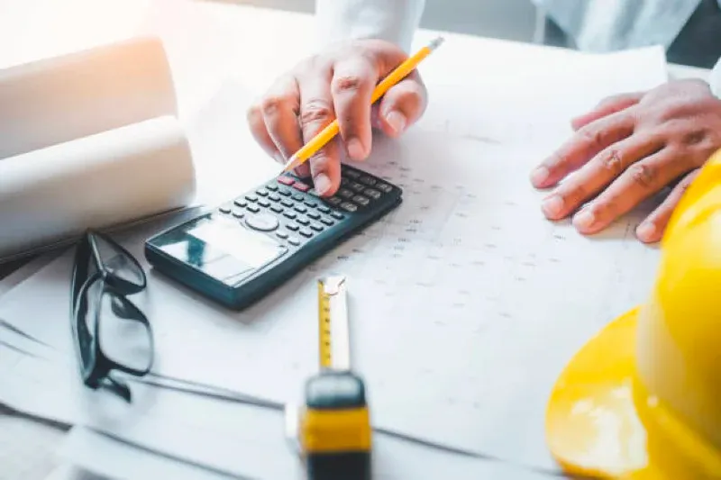 Hands using a calculator with pencil, tape measure, and hard hat on a desk
