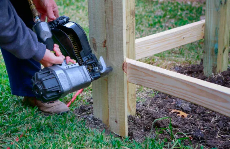 Person using a nail gun to attach wooden rails to a fence post outdoors