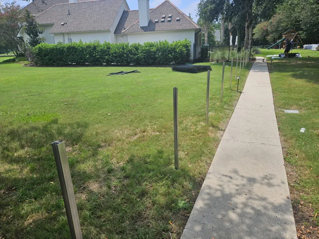 Residential lawn beside a sidewalk, with wooden stakes and houses in the background.