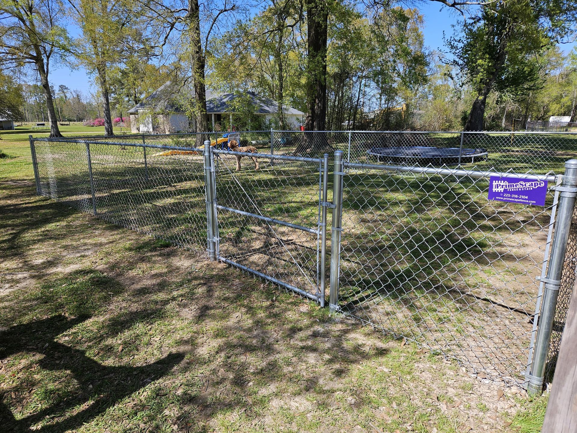 Chain-link gate in a grassy park, with trees and a small purple sign on the fence.