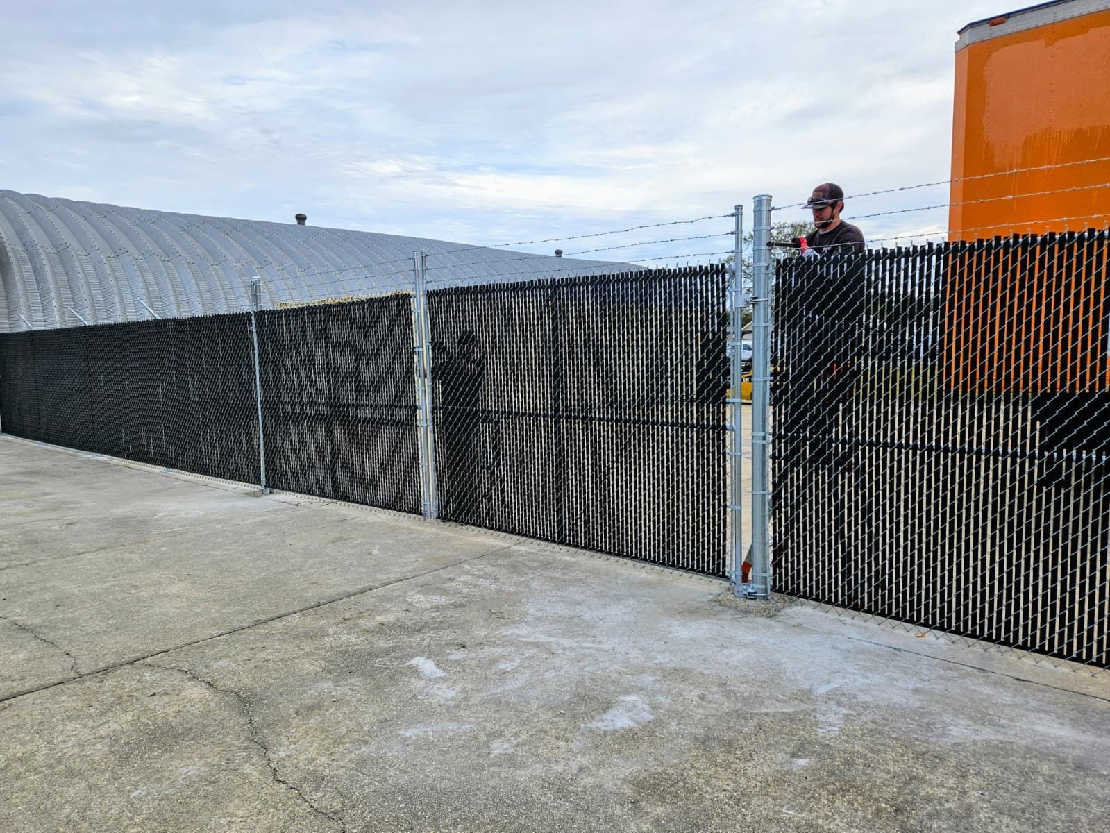Industrial yard with black mesh fencing, orange tank, and a person standing near the gate