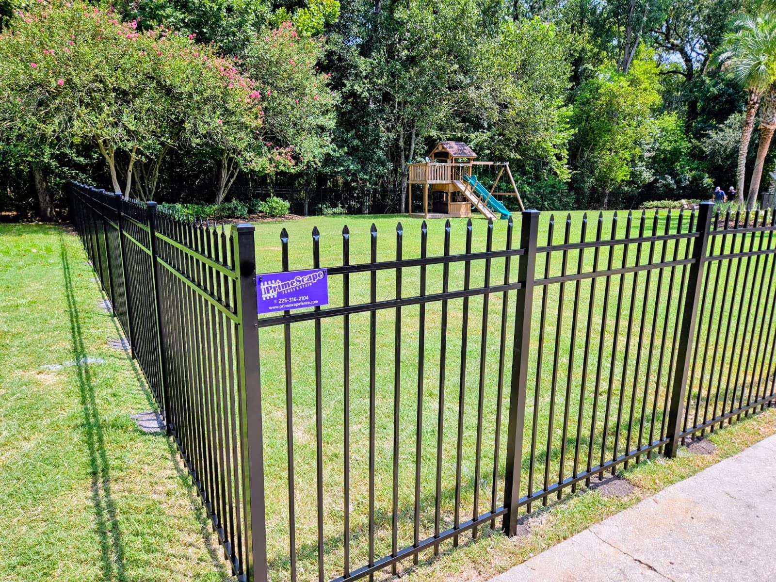 Black metal fence beside a grassy yard with trees and a purple sign