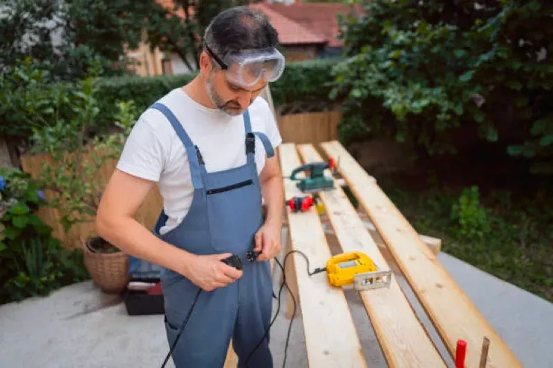 Man wearing safety goggles and overalls working with tools on a wooden workbench outdoors