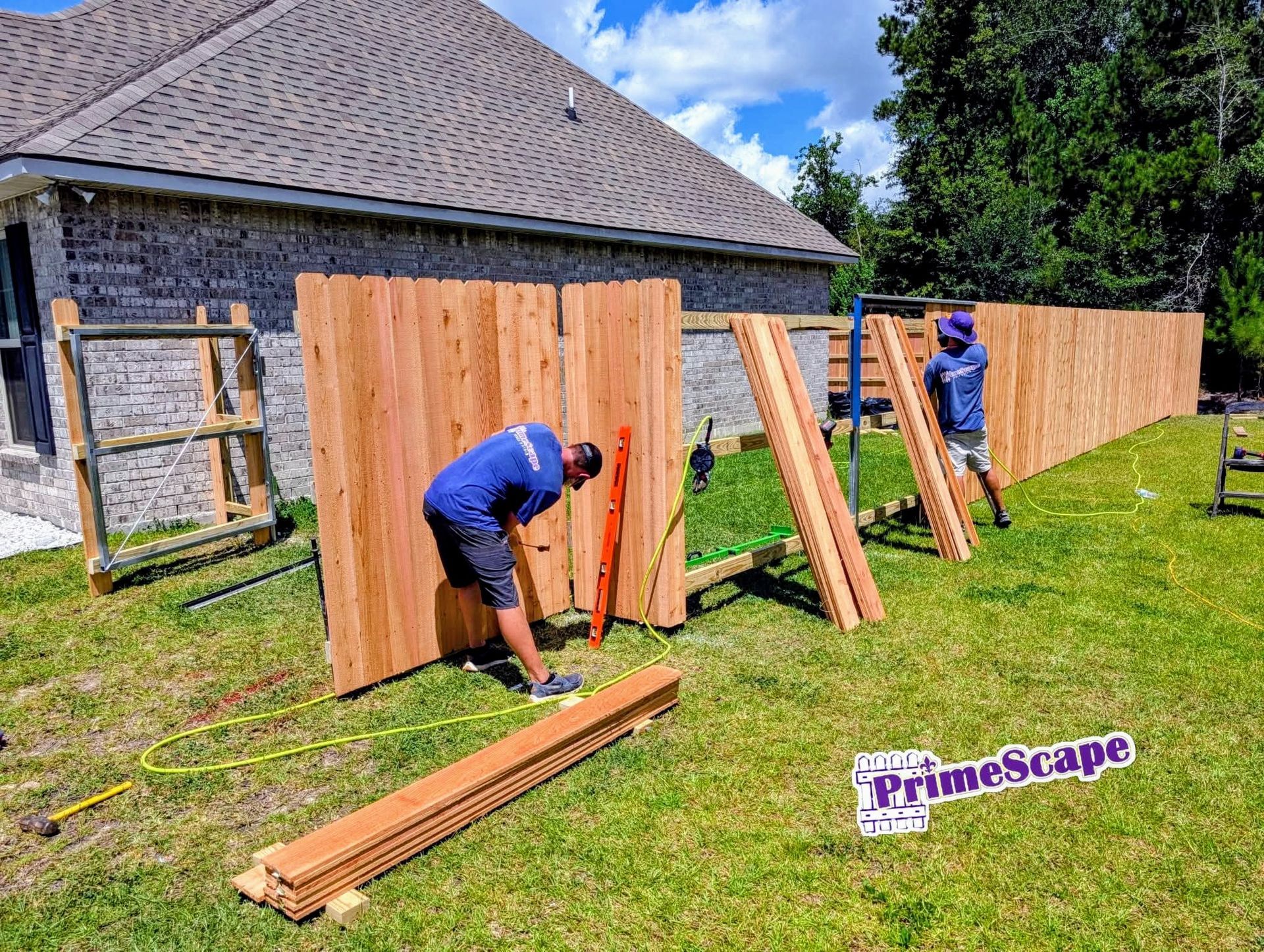 Two workers install a wooden fence in a grassy yard beside a house.