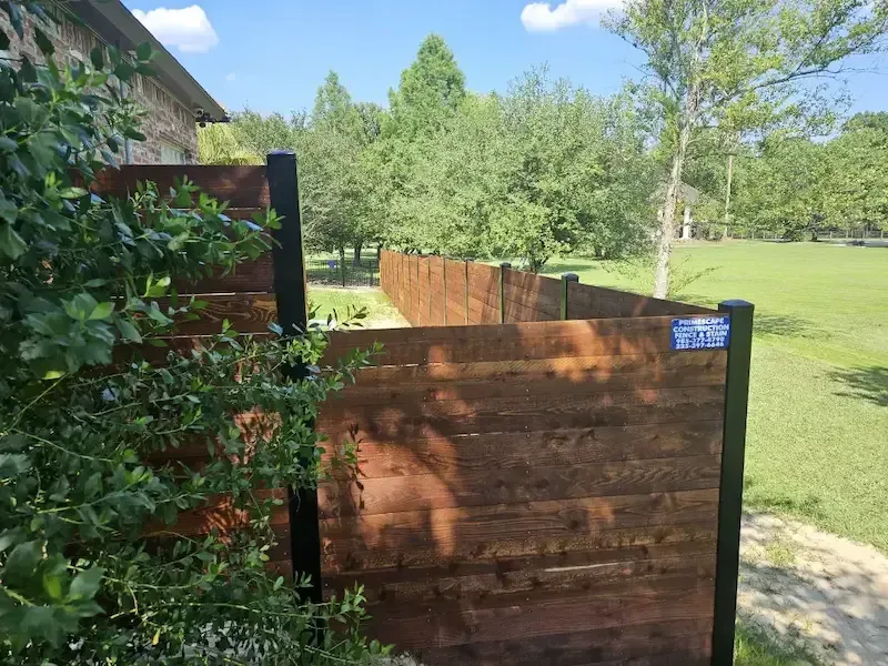 Wooden gate beside a leafy hedge, opening to a grassy park under a bright blue sky.