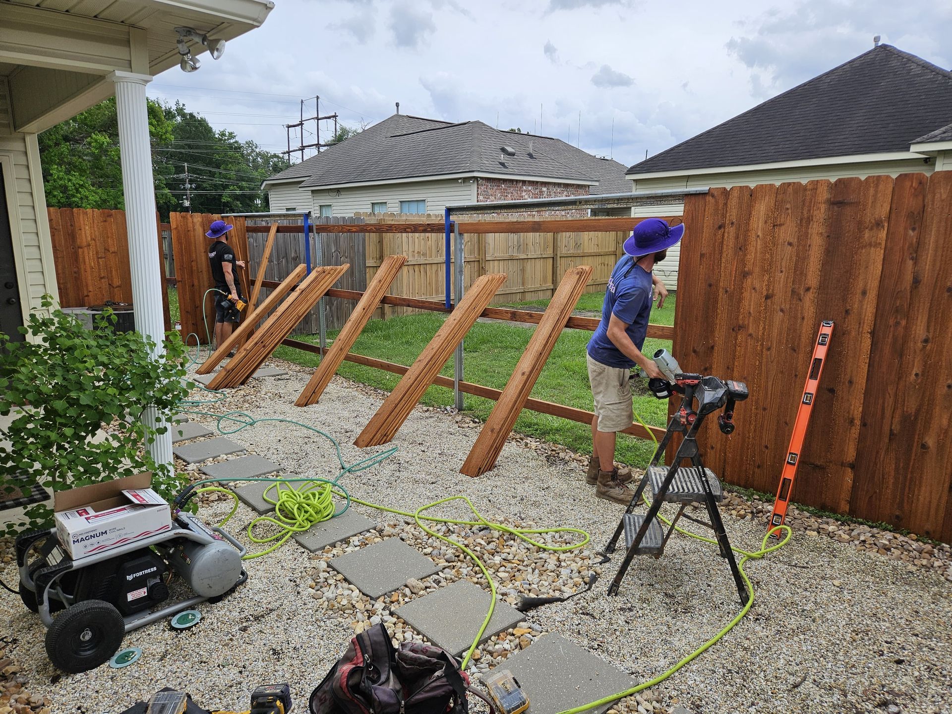 Person spraying a wooden fence in a sunny backyard with green lawn and patio.