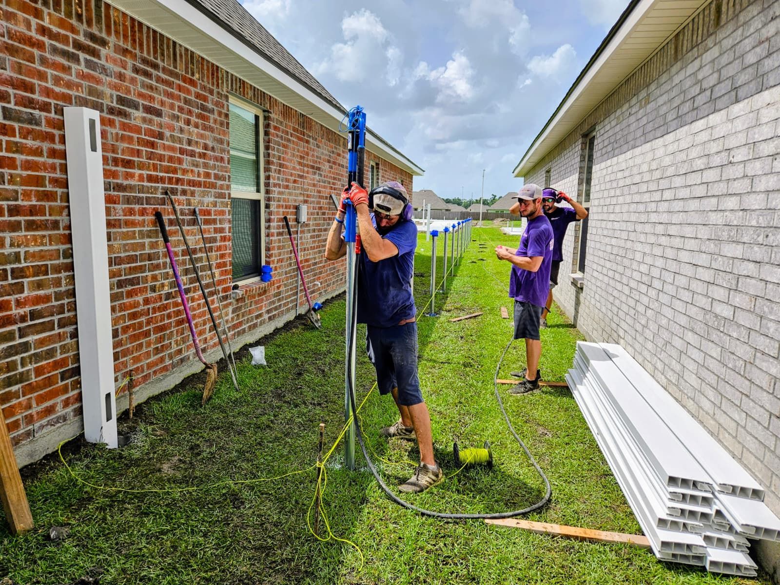 Two workers setting up equipment between a brick house and white siding, with ladders and hoses on the grass.