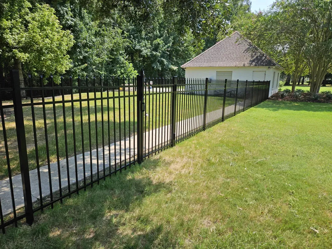 Black metal fence beside a grassy yard, with trees and a small white building in the background