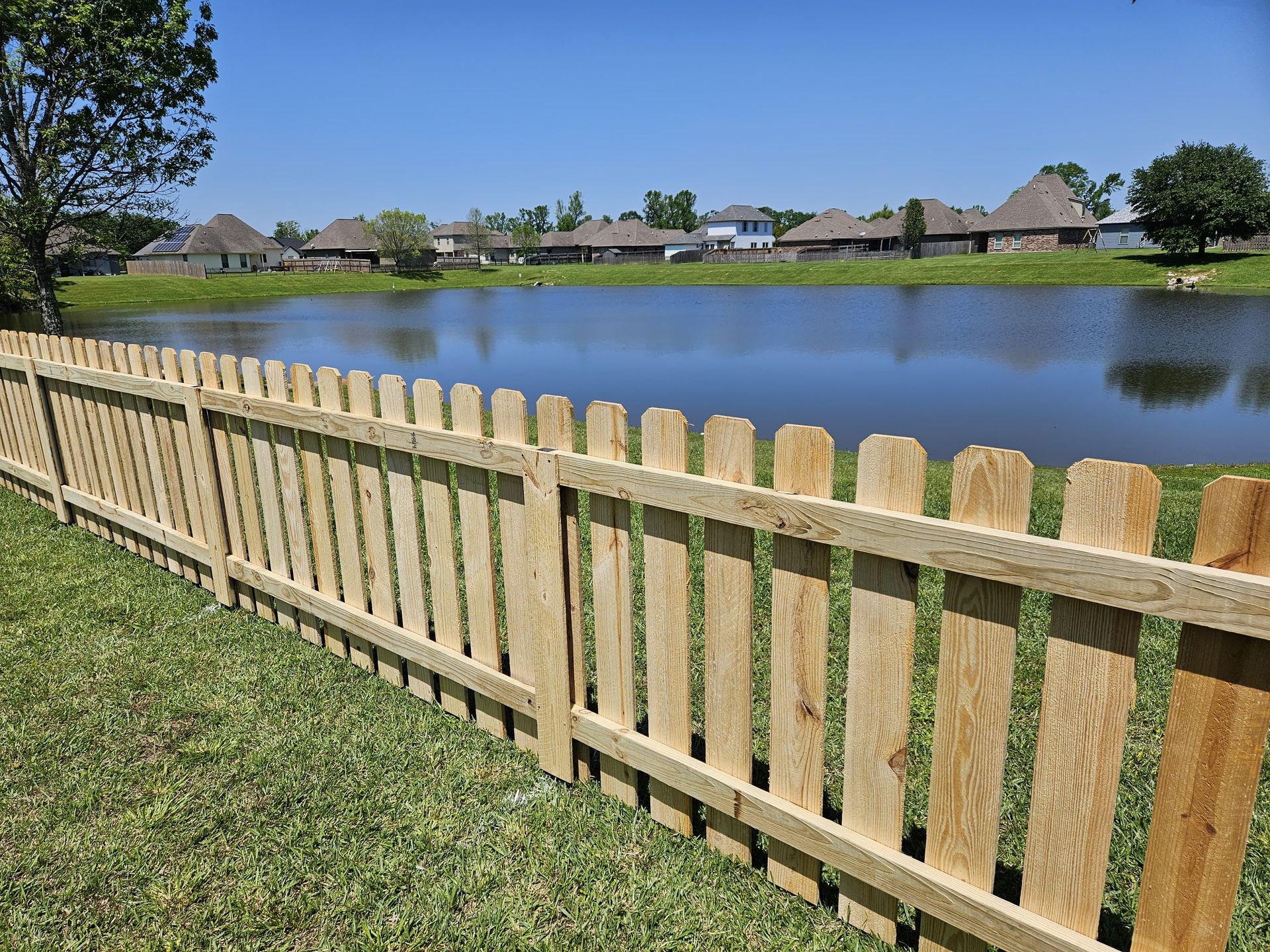 Wooden fence beside a calm lake in a grassy suburban neighborhood under a clear blue sky