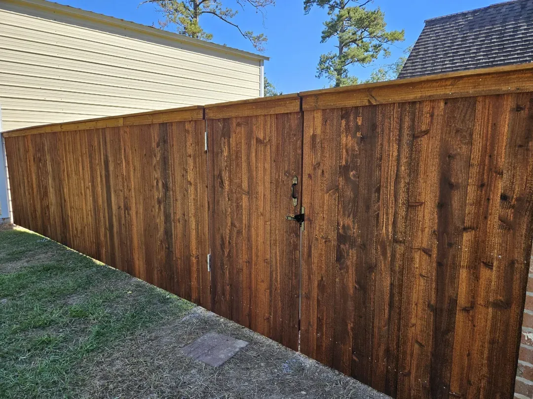 Wooden privacy fence with gate beside a beige house, next to a grassy yard under a blue sky.