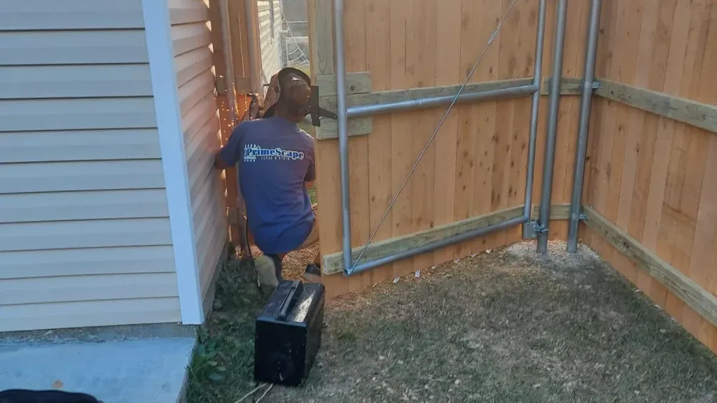 Person crouches by a wooden fence, placing a black suitcase in a narrow backyard corner.