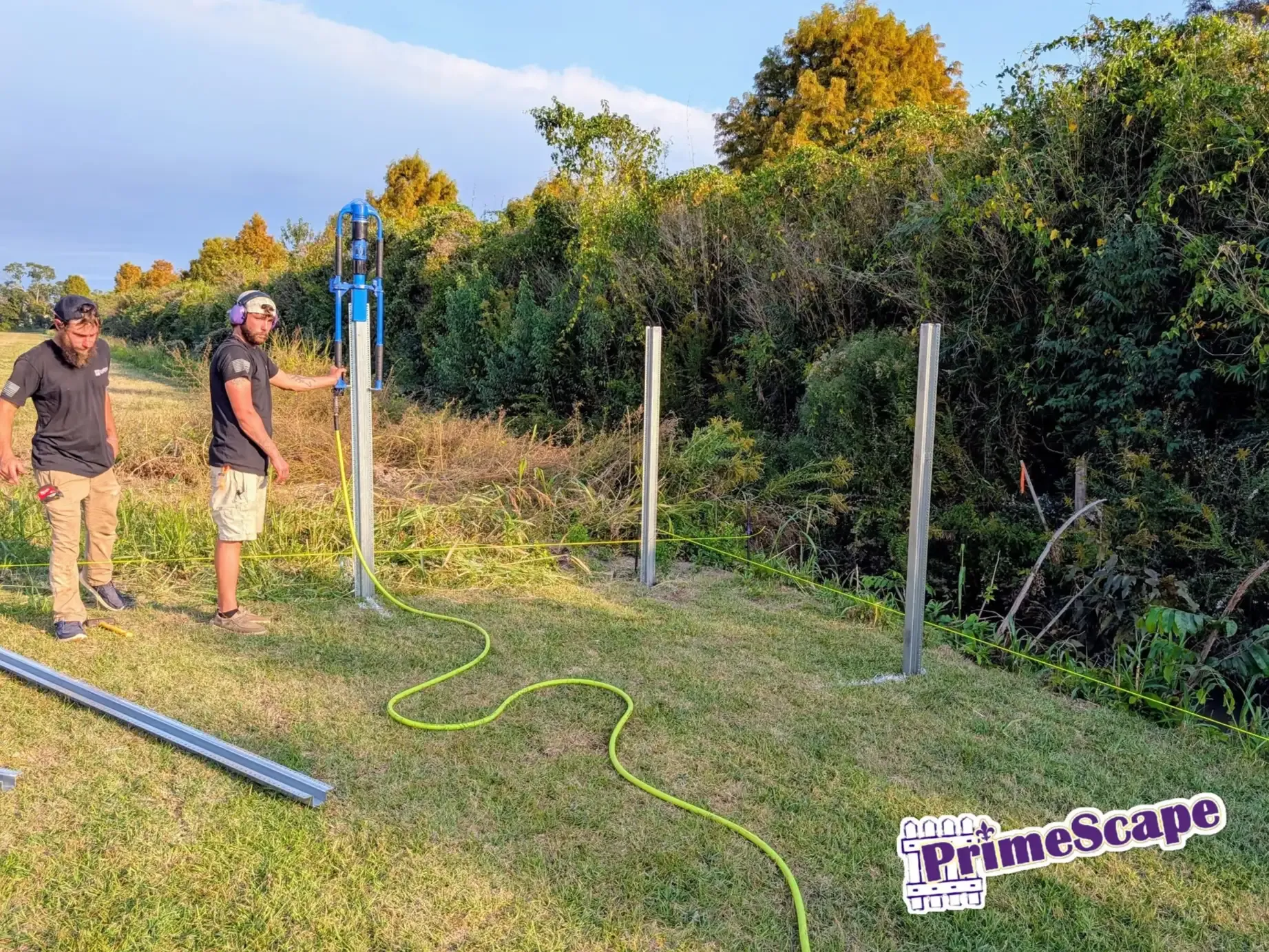 Two people standing beside fence posts in a grassy field with a green hose and blue sky.