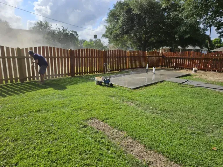 Person working by a wooden fence in a grassy backyard with smoke drifting in the background.