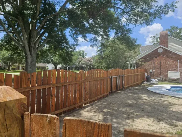 Wooden backyard fence beside a brick house, with trees and a small pool under a blue sky.