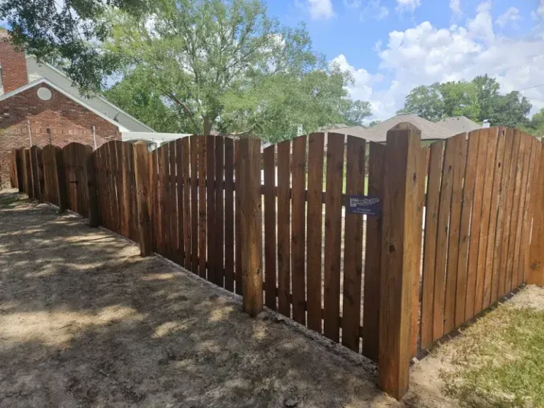 Wooden privacy fence along a dirt path beside a house under a blue sky.