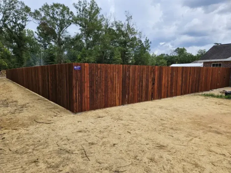 Newly built dark wood fence along a sandy yard with trees and a house in the background