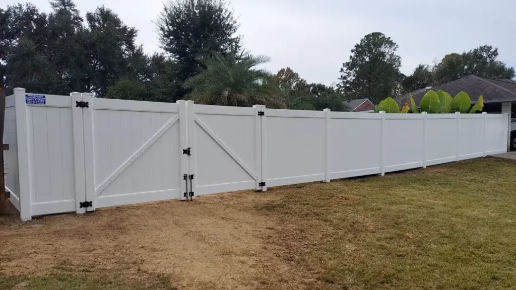 White vinyl fence with a double gate along a grassy yard, with trees and houses in the background