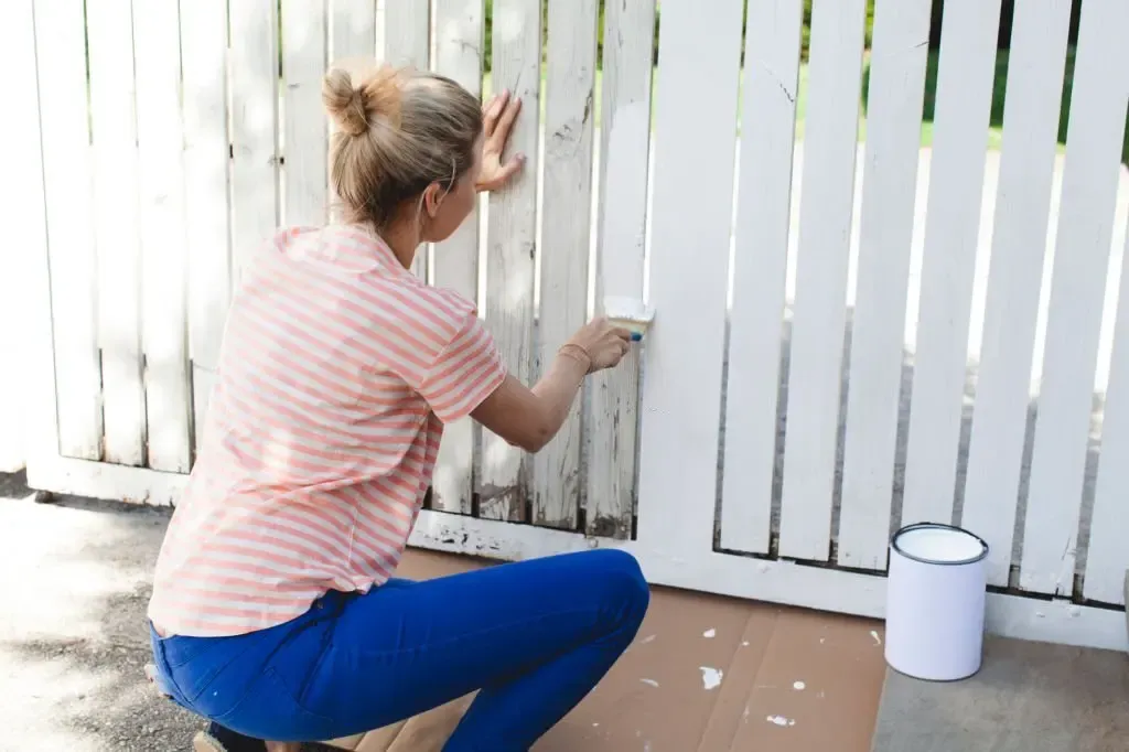 Person painting a white wooden fence outdoors with a small paint bucket nearby