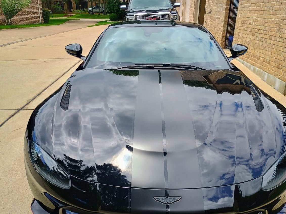 Black sports car parked in a driveway, viewed from the front with sky reflections on the hood.