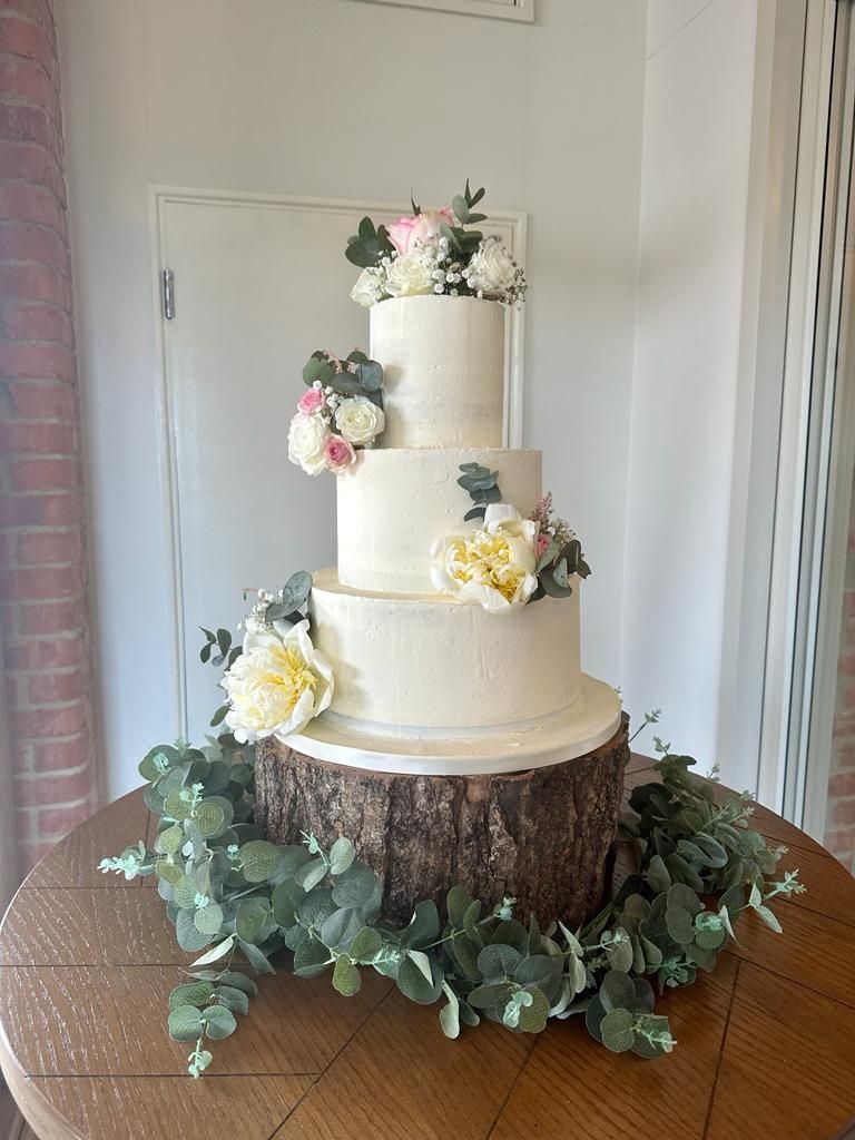 Four-tier white wedding cake with floral decorations on a rustic wood slice, surrounded by greenery