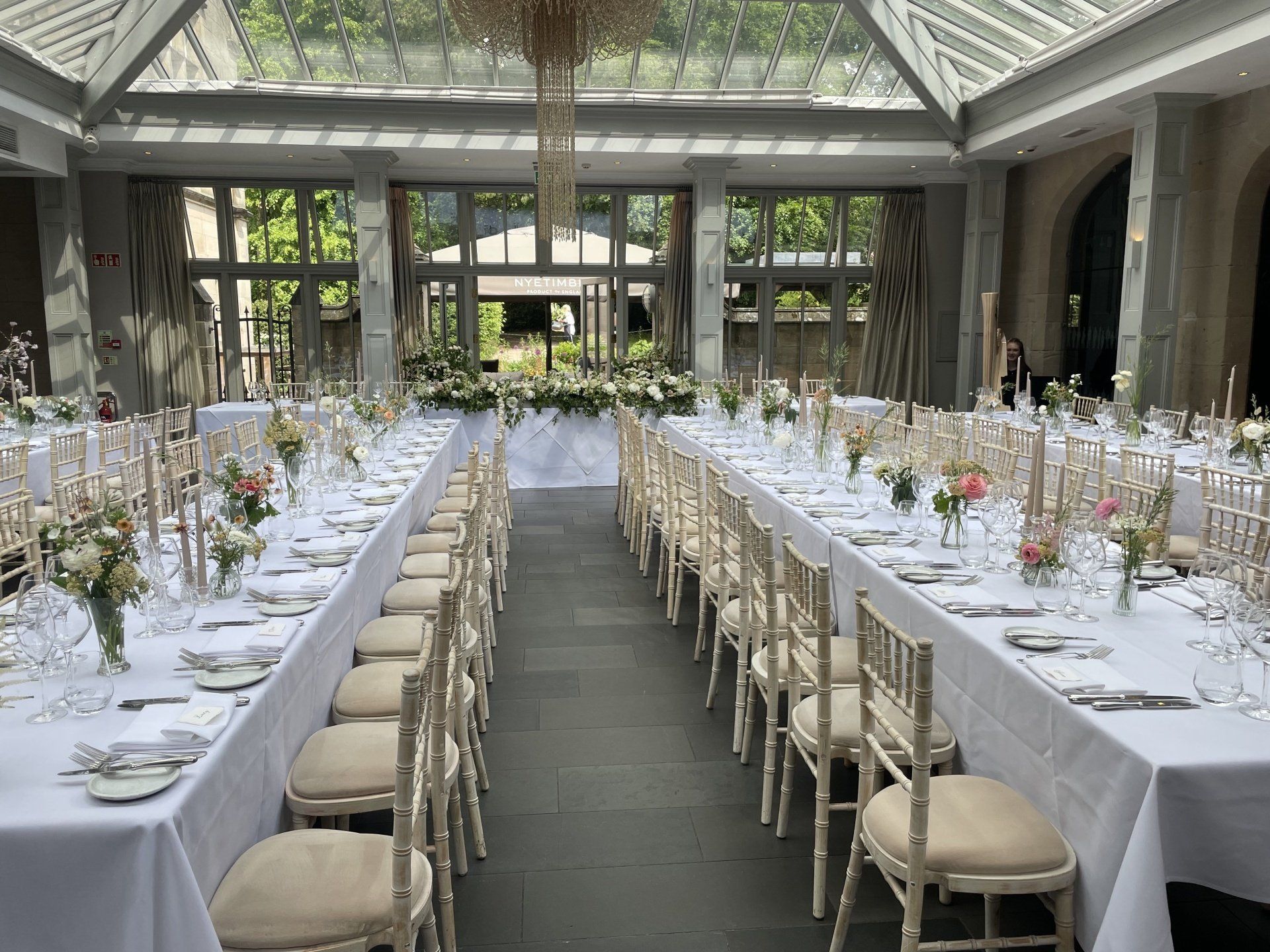 Elegant outdoor dining setup with long white-clothed tables and beige chairs in a glass-roofed patio.