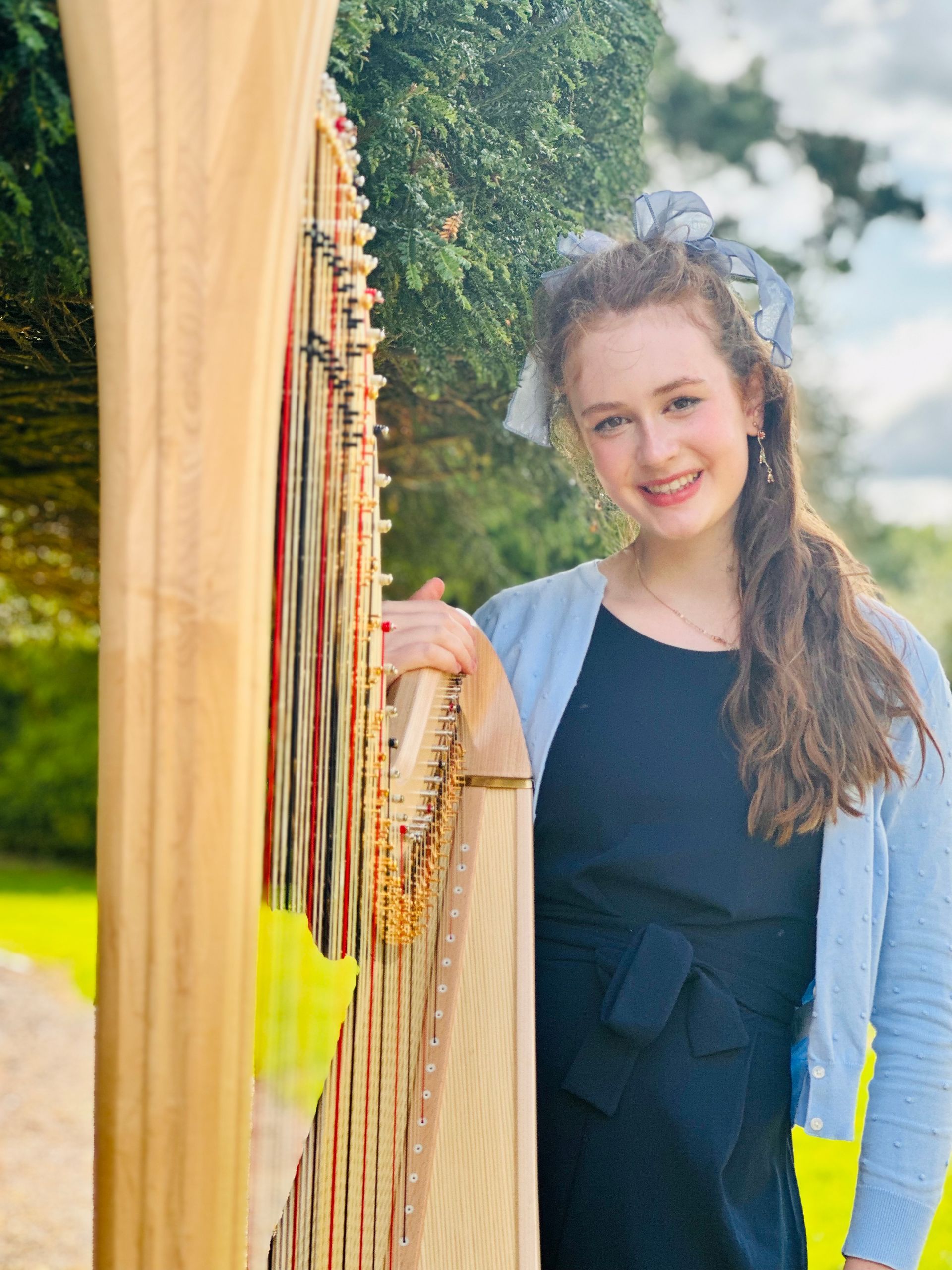 Sebrina smiling beside a harp outdoors, with trees and bright sunlight in the background