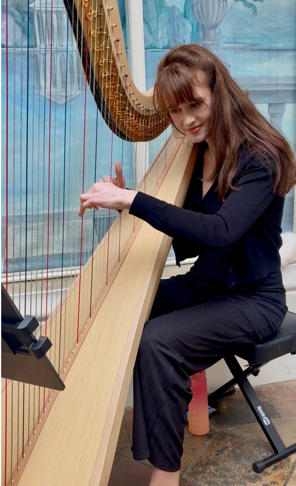 Sebrina in a black outfit playing a large harp indoors, with long hair falling forward.