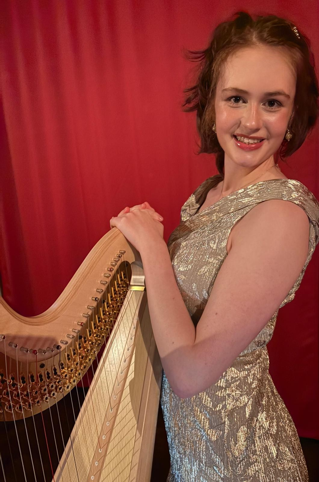 Smiling woman in a sparkling dress holding a harp against a red curtain backdrop