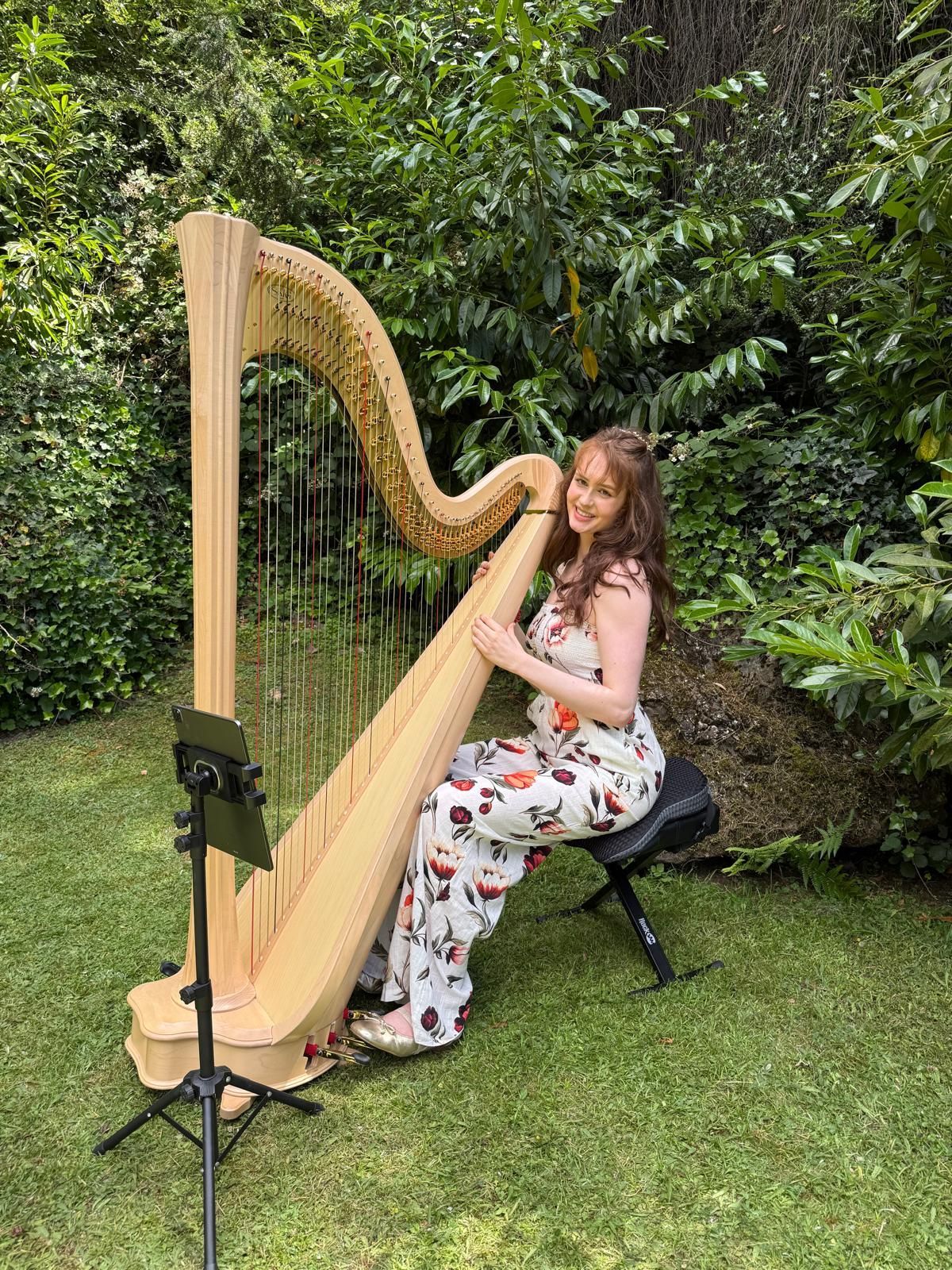 Woman playing a golden harp outdoors in a lush garden, seated on a black stool.