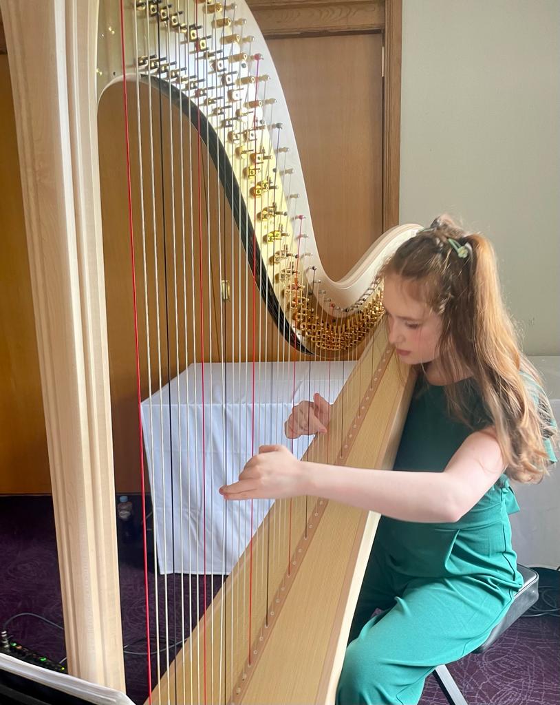 Sebrina in a green outfit playing a tall gold harp indoors near a window.
